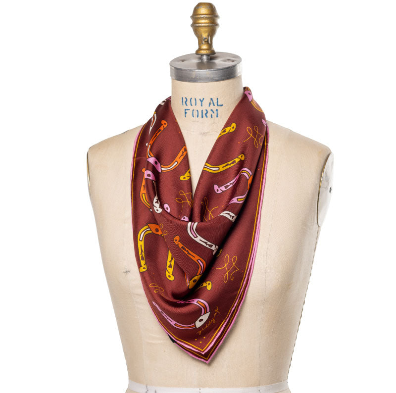 A maroon silk scarf with a pattern of horseshoes, keys, and musical notes, displayed on a mannequin.