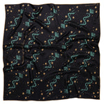 This image features the "Lasso the Stars" silk scarf, elegantly displayed on a wooden hanger. The scarf showcases a deep midnight blue color adorned with whimsical patterns of teal serpents, gold stars, and floral accents.