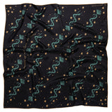 This image features the "Lasso the Stars" silk scarf, elegantly displayed on a wooden hanger. The scarf showcases a deep midnight blue color adorned with whimsical patterns of teal serpents, gold stars, and floral accents.