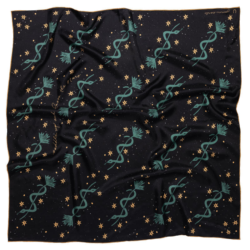 This image features the "Lasso the Stars" silk scarf, elegantly displayed on a wooden hanger. The scarf showcases a deep midnight blue color adorned with whimsical patterns of teal serpents, gold stars, and floral accents.
