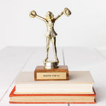 A trophy featuring a metal cheerleader figurine with arms raised in celebration, mounted on a wooden base with the inscription 'ROOTIN FOR YA'.
