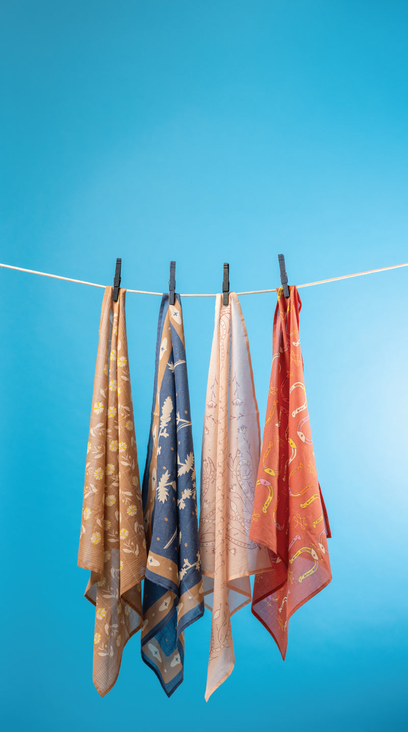 Four patterned silk scarves in beige, blue, cream, and rust orange hang from a white clothesline with black clips against a bright blue gradient background.