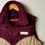 A color-blocked puffer vest hanging on a wooden hanger against a plain light wall. The vest has a beige lower body and a dark burgundy upper yoke, collar, and zipper trim.