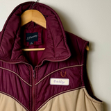 A color-blocked puffer vest hanging on a wooden hanger against a plain light wall. The vest has a beige lower body and a dark burgundy upper yoke, collar, and zipper trim.