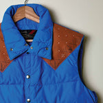 A blue puffer vest with brown geometric-patterned shoulder panels, hanging on a wooden hanger against a light gray wall.