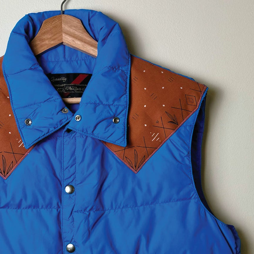 A blue puffer vest with brown geometric-patterned shoulder panels, hanging on a wooden hanger against a light gray wall.