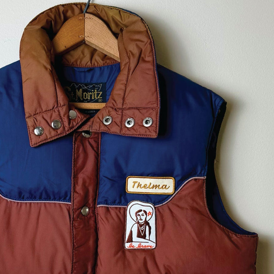 A vintage colorblock puffer vest hanging on a wooden hanger against a plain light gray wall. The vest features a dark brown body, a royal blue upper yoke and collar, and silver metal snap buttons and zippers.