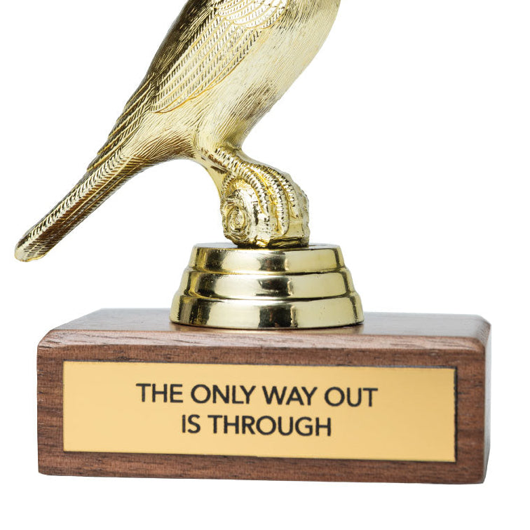 A gold bird figurine perched on a small gold base, mounted on a rectangular brown wooden plaque with a gold nameplate that reads 'THE ONLY WAY OUT IS THROUGH' on a white background.