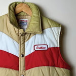 A vintage quilted puffer vest in a colorblock pattern of beige, white, and red, hanging on a wooden hanger against a plain light gray wall. The vest features a beige lower body, a white upper chest, and a red band across the chest.