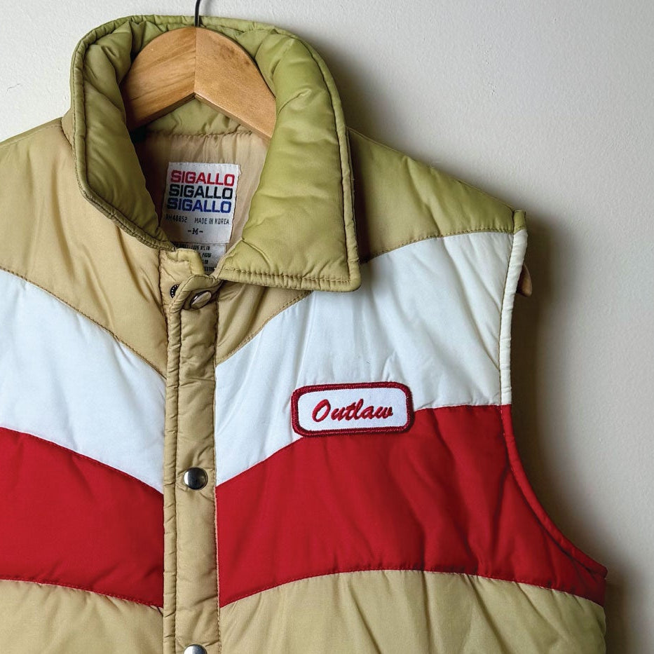 A vintage quilted puffer vest in a colorblock pattern of beige, white, and red, hanging on a wooden hanger against a plain light gray wall. The vest features a beige lower body, a white upper chest, and a red band across the chest.