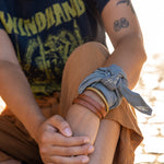 this is our "roam free" hand dyed bandana worn tied around the wrist with some bangle bracelets, with the mantra "roam free" in the corner. the base color is lichen with light tan ink. there is a mountain and hatch mark pattern throughout the bandana.